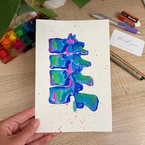May include: Watercolor painting of a spine in shades of blue, green, and pink on white paper. The artwork features a detailed anatomical representation with colorful paint splatters. A small card with the words "Thank you" is also visible.