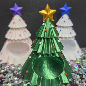 May include: Three decorative Christmas tree ornaments. The front tree is green with a gold star, red and white ornaments. Two white trees with purple and blue stars and red and blue ornaments are behind it. The ornaments are on a glittery surface.