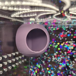 May include: A lavender-colored, spherical pet habitat with a large circular opening. The habitat is made of a textured material and is mounted inside a clear enclosure. The background features a colorful, glittery pattern.