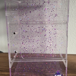 Op de afbeelding: Een heldere acryl display case met een paars glitter ontwerp. De case heeft een rechthoekige vorm met een scharnierende deur en een klein logo in de rechter benedenhoek. De glitter is verspreid over de case.