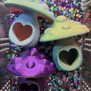 May include: Four colorful mushroom-shaped objects with heart-shaped cutouts. The mushrooms are in shades of blue, green, and purple. The caps have a textured, dotted surface. The background is a sparkly, iridescent pattern.