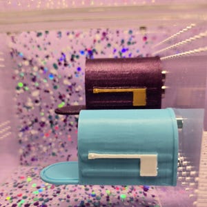 May include: Two miniature mailbox ornaments, one dark purple with a gold flag and one light blue with a white flag. Each mailbox features a rounded top, a small door, and a side-mounted flag mechanism, presented in a clear display case.