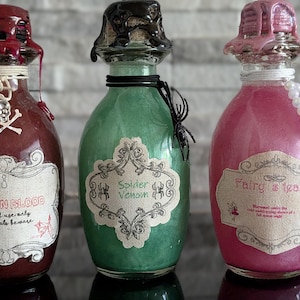 Può includere: Tre bottiglie decorative di pozioni con etichette che recitano "Dragon Blood", "Spider Venom" e "Fairy Tears". Le bottiglie sono riempite con liquidi colorati e decorate con tappi decorativi, ciondoli a forma di teschio e una collana di perle.