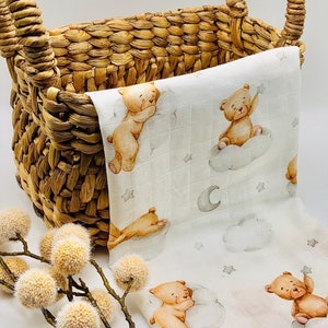 May include: A white muslin baby blanket with a repeating pattern of brown teddy bears, clouds, and stars. The blanket is folded and placed in a woven basket with handles.