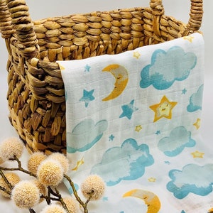 May include: A white muslin baby blanket with a blue and yellow moon, stars, and clouds pattern. The blanket is folded and placed in a woven wicker basket with handles.