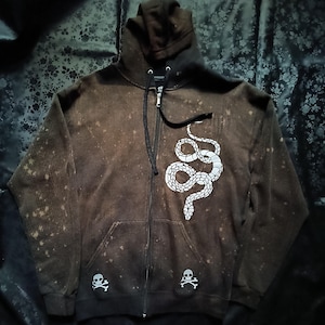 May include: Brown zip-up hoodie with a white snake design on the front. The hoodie has a distressed look with a black zipper and two small skull and crossbones graphics near the pockets. The hood is dark brown.