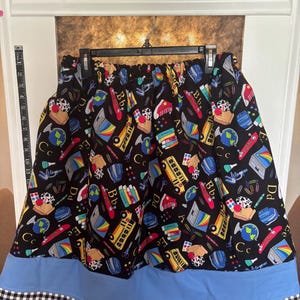 Pode incluir: Uma saia preta com uma estampa escolar colorida de autocarros amarelos, globos, livros e letras. A saia tem uma faixa larga azul-clara na bainha, com um acabamento em xadrez preto e branco. Uma fita métrica é visível no lado esquerdo.