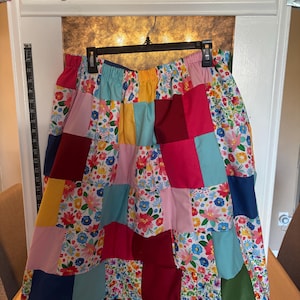 Op de afbeelding: Een patchwork rok met een verscheidenheid aan kleurrijke stoffen vierkanten. De rok heeft bloempatronen, effen kleuren en een comfortabele elastische tailleband. De bovenkant van het display leest "ELKM" met een schaar en een speldenkussen.