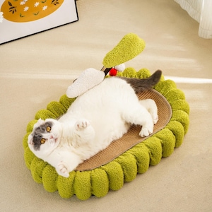 May include: A white and gray cat laying in a green oval-shaped cat bed with a scratching post and a plush toy attached. The bed is made of soft, fluffy material and has a textured surface.
