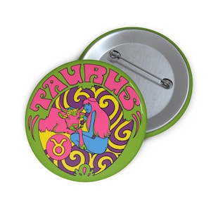 Puede incluir: Un botón verde con la palabra "TAURUS" en rosa y un diseño psicodélico. El botón presenta un toro rosa, una figura azul y el símbolo del zodiaco Tauro. Un segundo botón es visible en el fondo.