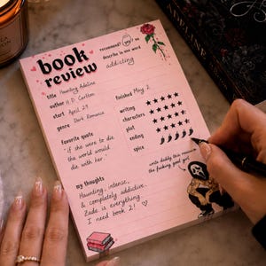 K&ouml;nnte beinhalten: Ein rosa Notizblock f&uuml;r eine Buchrezension mit dem Titel "book review" in schwarzer Schrift. Der Notizblock enth&auml;lt Abschnitte f&uuml;r Titel, Autor, Startdatum, Genre und Lieblingszitat. Eine Hand schreibt mit einem schwarzen Stift auf den Notizblock.