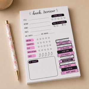 May include: A white book review notepad with black text and pink accents, featuring fields for title, author, genre, and ratings. Beside it lies a retractable pen with a pink floral pattern. The notepad includes a stack of illustrated books.