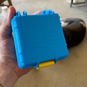 Könnte beinhalten: Eine blaue, rechteckige Box mit dem Text "VAULT-TEC" und einem Logo. Die Box hat einen gelben Verschluss und blaue Verschlüsse. Die Box wird in der Hand einer Person gehalten.