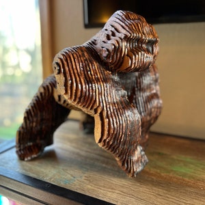 Può includere: Una scultura in legno di un gorilla, realizzata in legno stratificato, con una finitura marrone scuro. Il gorilla è in piedi sulle sue zampe posteriori, con le braccia distese.
