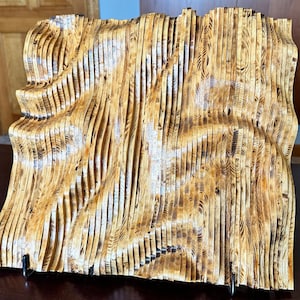 May include: A wall sculpture made of thin strips of wood, arranged in a wavy pattern. The wood is a light brown colour with darker brown streaks. The sculpture is mounted on a black stand.