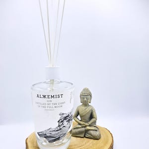 Puede incluir: Una botella de cristal transparente de ginebra Alxemist con varillas difusoras blancas, con una etiqueta que dice Alxemist Gin Distilled by the Light of the Full Moon, junto a una pequeña figura de Buda gris sobre una rodaja de madera.