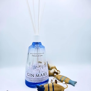 Puede incluir: Una botella de cristal Gin Mare con base en degradado azul, tapón blanco y cinco varillas difusoras blancas. La etiqueta dice Mediterranean Gin, Gin Mare, distilled with olives, thyme, rosemary and basil, 700ml, 42.7% Vol. Hay adornos de peces de madera cerca.