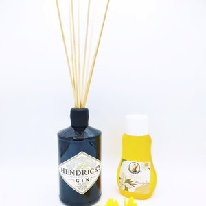 Könnte beinhalten: Eine schwarze Glasflasche mit schwarzem Verschluss und einem weißen Etikett, auf dem "Hendrick's Gin" mit einem kleinen gelben Vogellogo steht. Die Flasche ist mit einer klaren Flüssigkeit gefüllt und hat Holzstäbchen, die aus der Oberseite herausragen. Neben der größeren Flasche steht eine kleinere Flasche mit weißem Verschluss und gelber Flüssigkeit. Die kleinere Flasche hat ein weißes Etikett mit einem gelben Vogellogo und dem Text "Vanilla" in Schwarz. Zwei gelbe Sonnenblumen befinden sich im Vordergrund.