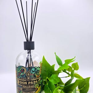 Può includere: Una bottiglia in vetro trasparente riutilizzata come diffusore a bastoncini con un tappo nero e sei bastoncini neri. L'etichetta riporta il testo Don Papa Masskara con intricate illustrazioni verdi, dorate e rosse di fogliame e una farfalla.