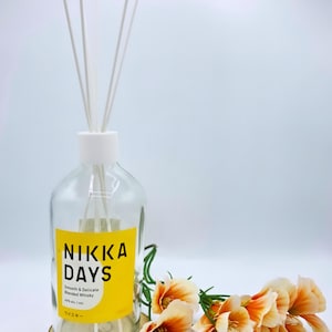 Può includere: Una bottiglia in vetro trasparente riutilizzata come diffusore a bastoncini con un tappo bianco e quattro bastoncini bianchi. L'etichetta gialla presenta il testo nero NIKKA DAYS, Smooth & Delicate Blended Whisky, 40% alc./vol. e caratteri giapponesi. Fiori artificiali arancioni sono posizionati accanto alla base.