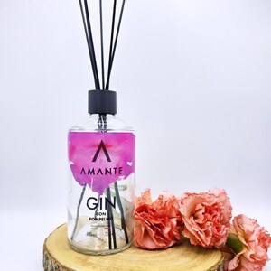 Könnte beinhalten: Eine durchsichtige Glasflasche für einen Raumduft mit schwarzem Verschluss und schwarzen Stäbchen. Das Etikett zeigt ein rosa Aquarellmuster, den Text AMANTE, GIN CON POMPELMO und PRODOTTO IN ITALIA. Die Flasche steht auf einer runden Holzscheibe.
