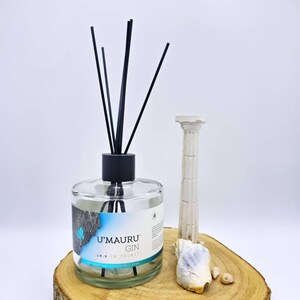 Puede incluir: Un difusor de varillas de vidrio transparente con tapa negra y varillas negras. La etiqueta presenta el texto U'MAURU GIN 19.9 IN SPIRIT con un gráfico azul y rocoso. Junto a la botella hay una pequeña figura de columna blanca y dos conchas.