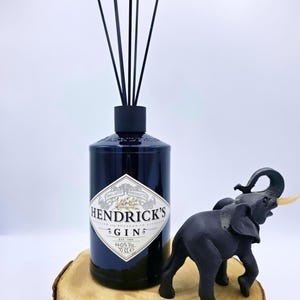 Può includere: Una bottiglia di gin Hendrick's blu scuro riutilizzata come diffusore a bastoncini neri, accompagnata da una piccola statuetta di elefante nero. L'etichetta riporta la scritta Hendrick's Gin, Est. 1886, 44.0% Vol. e 70 cl, su una fetta di legno.