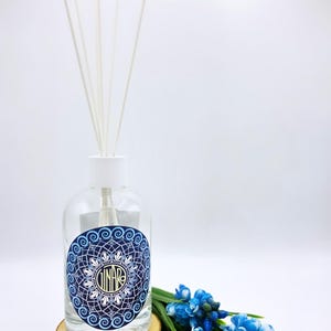 Può includere: Un flacone diffusore a bastoncini in vetro trasparente con tappo bianco e diversi bastoncini bianchi. Il flacone presenta un'etichetta blu circolare con un motivo mandala bianco e il testo GINARO al centro. Poggia su una fetta di legno rotonda.