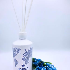 Può includere: Una bottiglia cilindrica bianca con una grafica blu di una mappa del mondo, il testo NORDÉS GIN THE TASTE OF ATLANTIC SPAIN e una rosa dei venti. Quattro sottili bastoncini diffusori bianchi sporgono dall'alto. Fiori blu accanto alla bottiglia.
