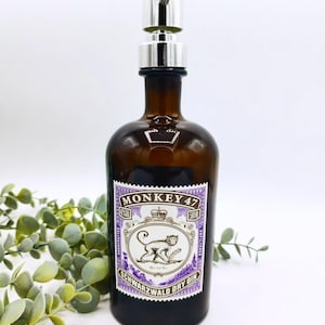 Könnte beinhalten: Eine braune Glasflasche mit einem silbernen Pumpspender. Die Flasche hat ein Etikett mit einem violetten Rand und dem Text "MONKEY 47 SCHWARZWALD DRY GIN".