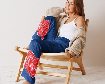 May include: A woman wearing a white vest top and a beige cardigan sits on a wooden chair. She is wearing blue satin trousers with a red floral pattern.