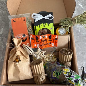 May include: A cardboard gift box filled with Halloween-themed items including a bag labelled Happy Halloween, a black box with green slime graphics, a bundle of dried grass, corrugated cardboard rolls, a star-shaped toy, and various small treats.