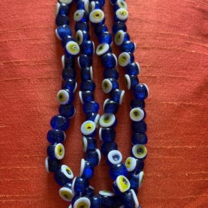 May include: A strand of blue glass beads with white and yellow evil eye charms. The beads are strung together in a long, continuous strand.