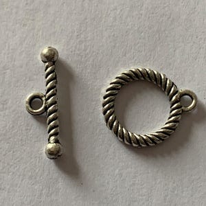 Może przedstawiać: Srebrny metalowy zamek z wzorem skręconej liny. Zamek ma okrągłą pętlę na jednym końcu i prosty pręt z okrągłą pętlą na drugim końcu.