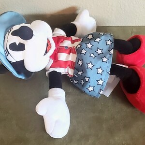 May include: A plush Minnie Mouse doll wearing a red and white striped top, a blue skirt with white stars, and a blue hat. The doll has black ears, a white face, and red shoes. The doll is lying on its back.
