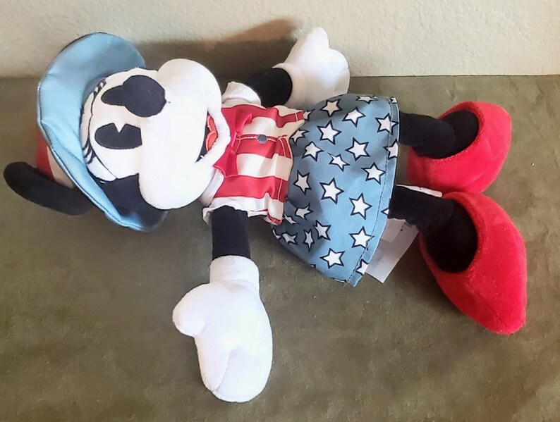 May include: A plush Minnie Mouse doll wearing a red and white striped top, a blue skirt with white stars, and a blue hat. The doll has black ears, a white face, and red shoes. The doll is lying on its back.