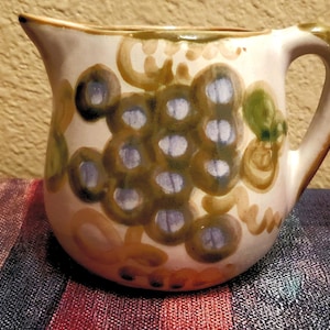 May include: A white ceramic pitcher with a brown handle. The pitcher is decorated with a hand-painted design of green grapes with purple highlights.