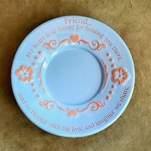 May include: A light blue ceramic saucer with a pink floral design and the text "Friend... My heart is so happy for holding you there. and so blessed with the love and laughter we share."