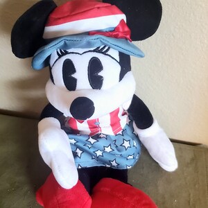 May include: A plush Minnie Mouse doll wearing a patriotic outfit. She has black ears, a red, white, and blue hat, and a dress with stars and stripes. Her shoes are red, and her arms and face are white.