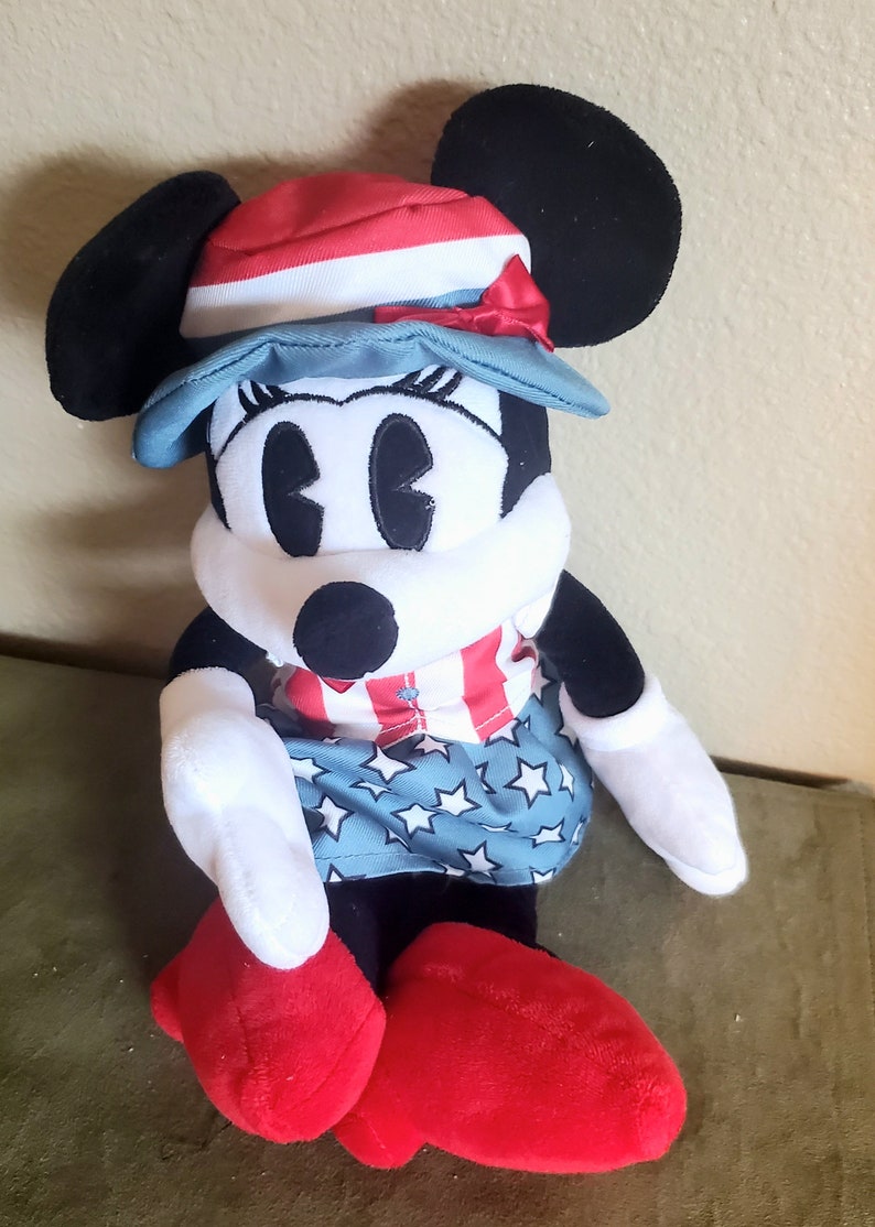 May include: A plush Minnie Mouse doll wearing a patriotic outfit. She has black ears, a red, white, and blue hat, and a dress with stars and stripes. Her shoes are red, and her arms and face are white.