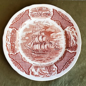 May include: A white ceramic plate with a red and white nautical design. The plate features a central image of a sailing ship, surrounded by decorative borders and text including "Friendship of Salem".