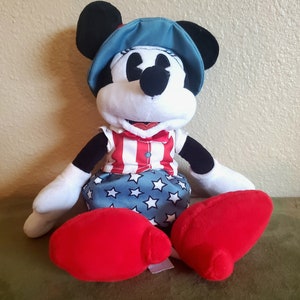 May include: A plush Minnie Mouse doll wearing a patriotic outfit. The doll has a blue hat, a red and white striped top, and a blue skirt with white stars. The doll's shoes are red.
