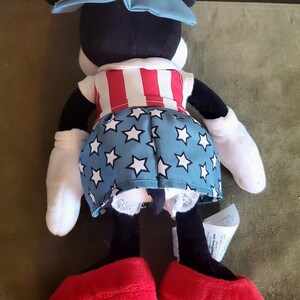 May include: A plush Minnie Mouse doll wearing a patriotic outfit. The doll has a red, white, and blue hat, a red and white striped top, and a blue skirt with white stars. It has large red shoes and white gloves.