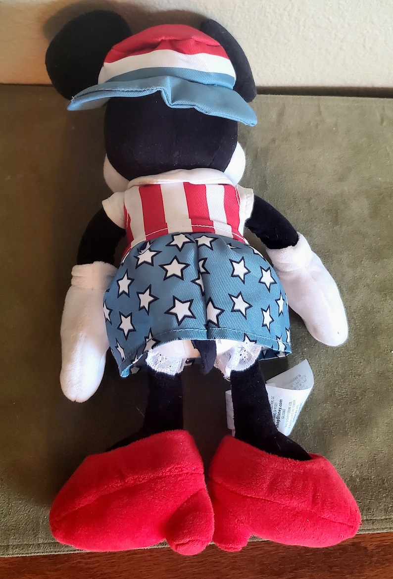 May include: A plush Minnie Mouse doll wearing a patriotic outfit. The doll has a red, white, and blue hat, a red and white striped top, and a blue skirt with white stars. It has large red shoes and white gloves.