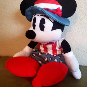May include: A plush Minnie Mouse doll wearing a patriotic outfit. She has a red, white, and blue hat, a red and white striped shirt, and blue shorts with white stars. Her large red shoes and black ears are prominent.