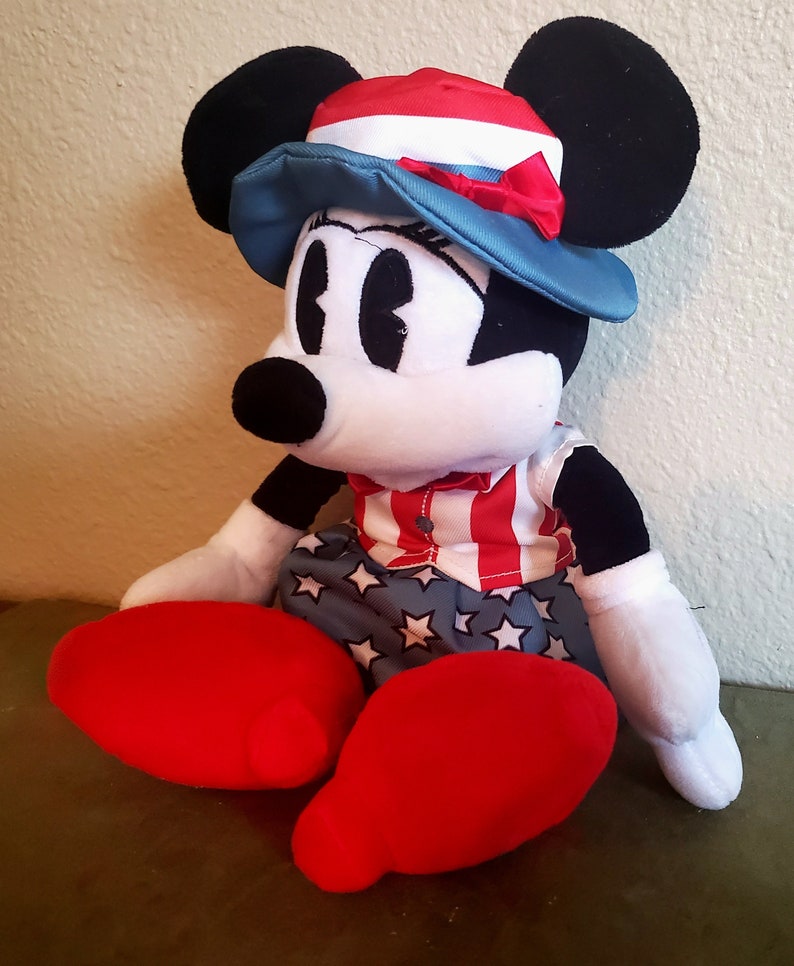 May include: A plush Minnie Mouse doll wearing a patriotic outfit. She has a red, white, and blue hat, a red and white striped shirt, and blue shorts with white stars. Her large red shoes and black ears are prominent.
