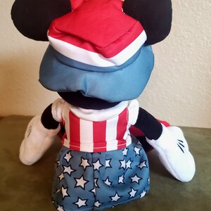 May include: A plush Mickey Mouse doll wearing a patriotic outfit. The doll has a red, white, and blue hat, a red and white striped shirt, and blue shorts with white stars. The doll has black ears, black arms, and white gloves.