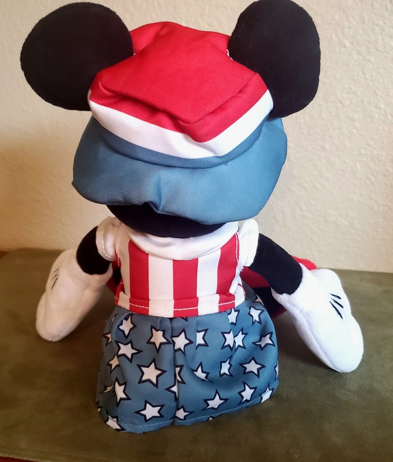 May include: A plush Mickey Mouse doll wearing a patriotic outfit. The doll has a red, white, and blue hat, a red and white striped shirt, and blue shorts with white stars. The doll has black ears, black arms, and white gloves.