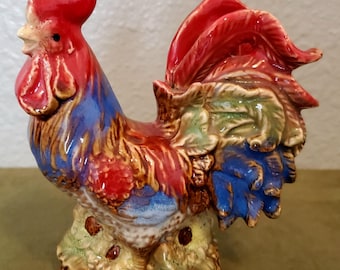 Pode incluir: Estatueta de galo de cerâmica com cores vibrantes. O galo apresenta crista e barbela vermelhas, penas azuis e vermelhas e uma base detalhada com detalhes verdes e castanhos. A estatueta é um objeto decorativo.