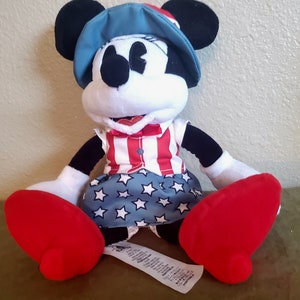 May include: A Minnie Mouse plush toy wearing a patriotic outfit. The toy has a blue hat, a red, white, and blue striped top, and a blue skirt with white stars. The plush has large red shoes and black ears.